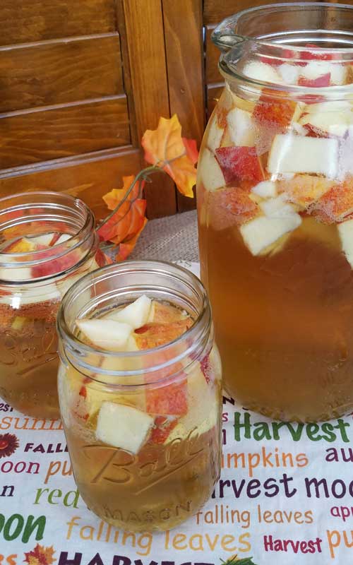 Two mason jars and a clear glass pitcher filled with Autumn Hard Apple Cider Sangria. The jars and pitcher also have chopped apples in the sangria.