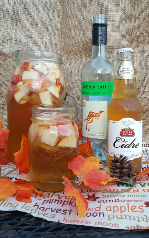 A mason jar and a clear glass pitcher filled with Autumn Hard Apple Cider Sangria. The jar and pitcher also have chopped apples in the sangria. To the right is a full bottle of hard apple cider, and an empty bottle of pinot grigio wine.