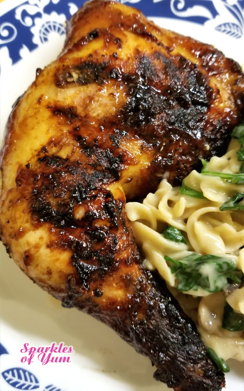 A grilled chicken leg quarter covered in barbecue sauce. The chicken is on a white plate, and resting next to noodles and spinach covered in a cream sauce.