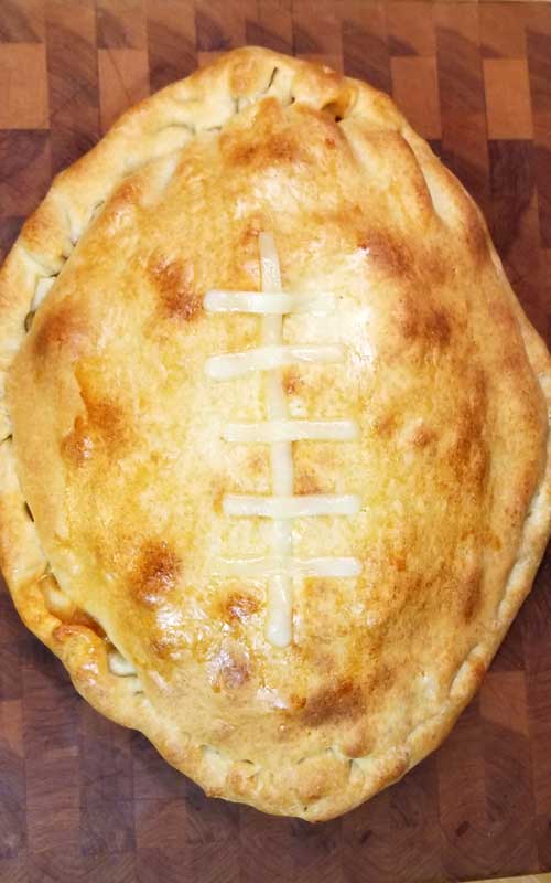 Top down view of a baked calzone on a wooden cutting board. Pieces of cheese are arranged to mimic laces on a football.