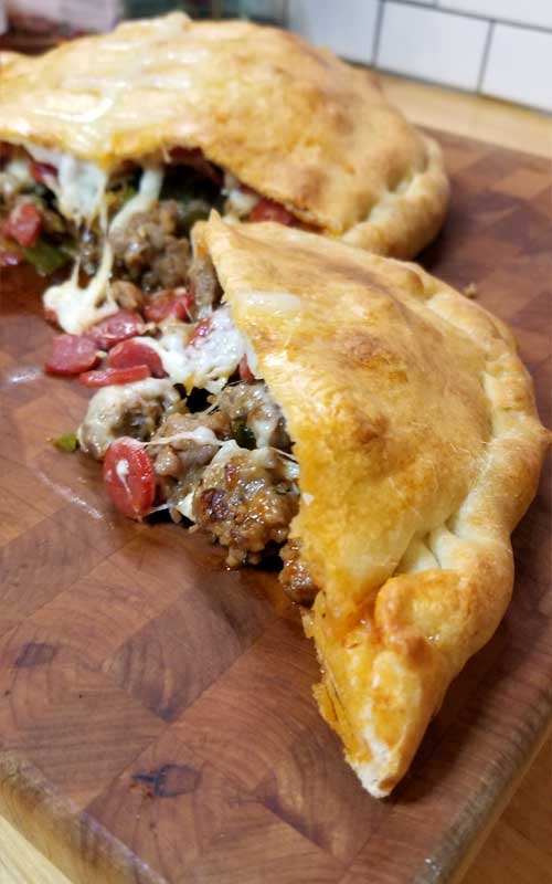 A split open calzone on a wooden cutting board. Sausage, pepperoni, and melted cheese are spilling out.