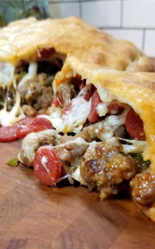 A split open calzone on a wooden cutting board. Sausage, pepperoni, and melted cheese are spilling out.