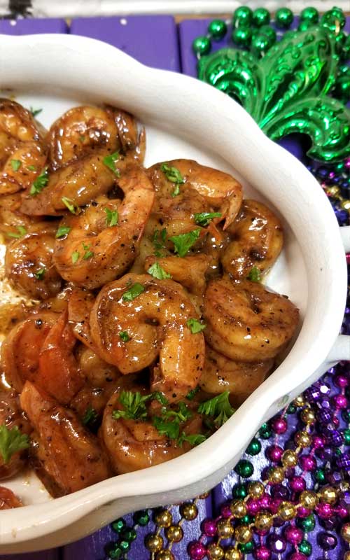 A top-down view of a white bowl containing New Orleans Barbeque Shrimp. The bowl is resting on gold, purple, and green Mardi Gras beads.