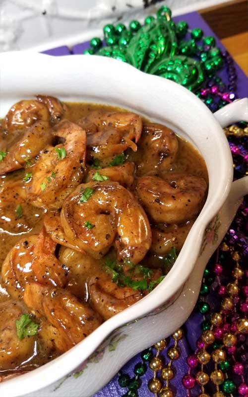 A white bowl containing New Orleans Barbeque Shrimp with extra gravy. The bowl is resting on gold, purple, and green Mardi Gras beads.