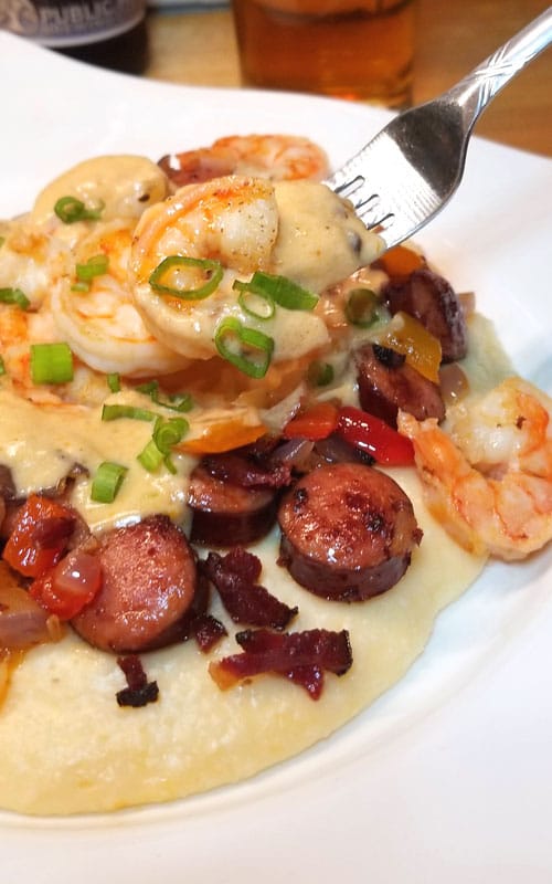 Shrimp and Grits being scooped up with a fork from a white plate . Grilled sausage, tasso ham, and green onion are also on the plate.