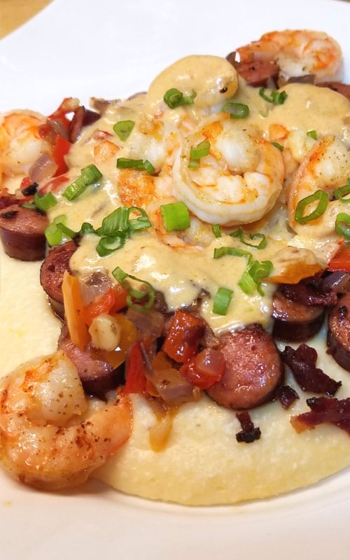 A close up view of Shrimp and Grits on a white plate. Cheese gravy, smoked sausage, and green onion cover the shrimp.