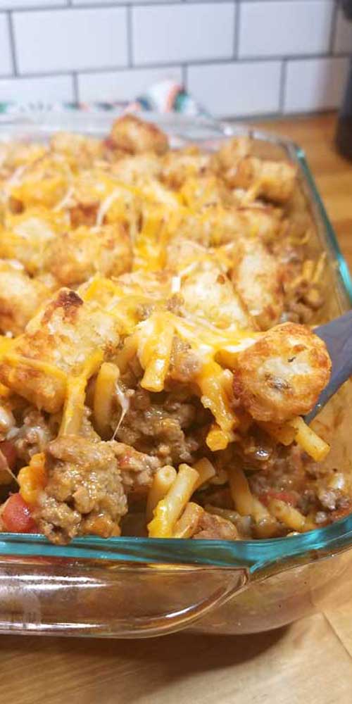 A spoon scooping out a serving of Mac n Cheesy Cowboy Casserole from a glass baking dish.