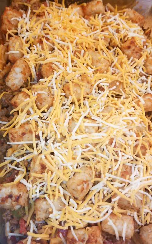An uncooked Mac n Cheesy Cowboy Casserole in a glass baking dish. The cheese and tater tots are clearly visible at the top.