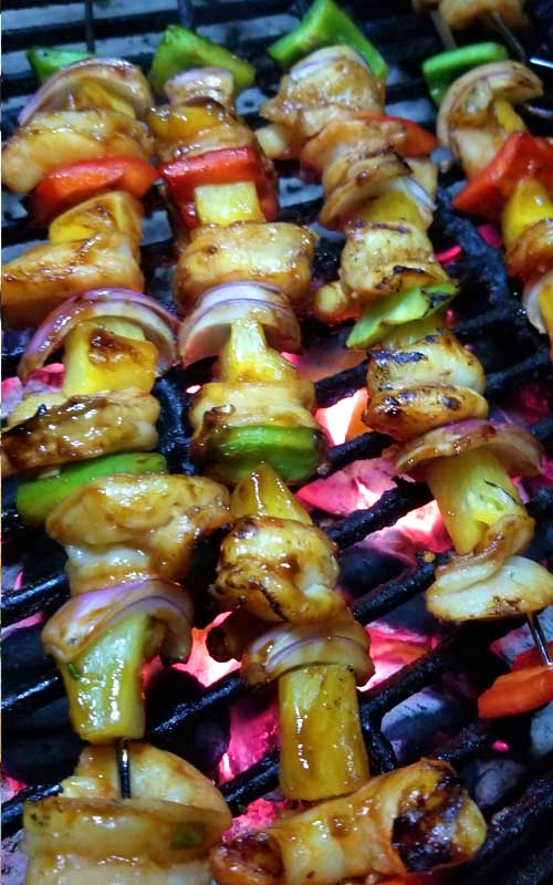 Chicken, bell peppers, onion, and pineapple on metal skewers being grilled on a cast iron grate.