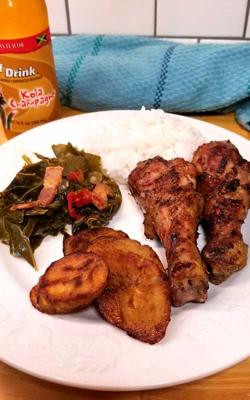 Two chicken legs, fried plantains, collard greens, and rice on a white plate. A bottled soft drink and blue towel are in the background.