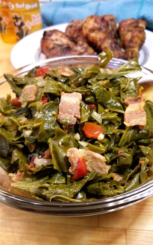 Very close up image of torn collard greens in a clear glass bowl. Pieces of bacon, onion, tomato, and garlic are scattered throughout the greens. In the background is a white plate topped with grilled chicken legs.