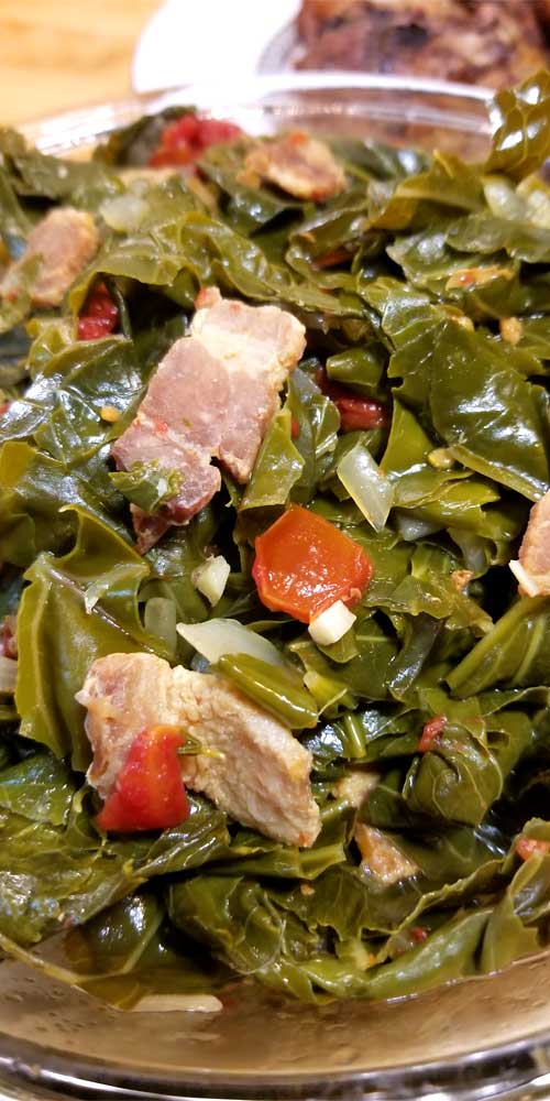Very close up image of torn collard greens in a clear glass bowl. Pieces of bacon, onion, tomato, and garlic are scattered throughout the greens.