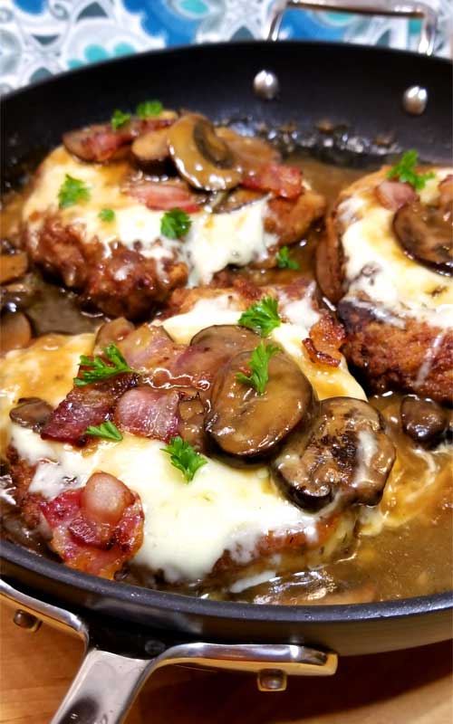 Three pork chops being cooked in a non-stick skillet. Each pork chop is covered with Fontina cheese, bacon, mushrooms, and a mushroom gravy.