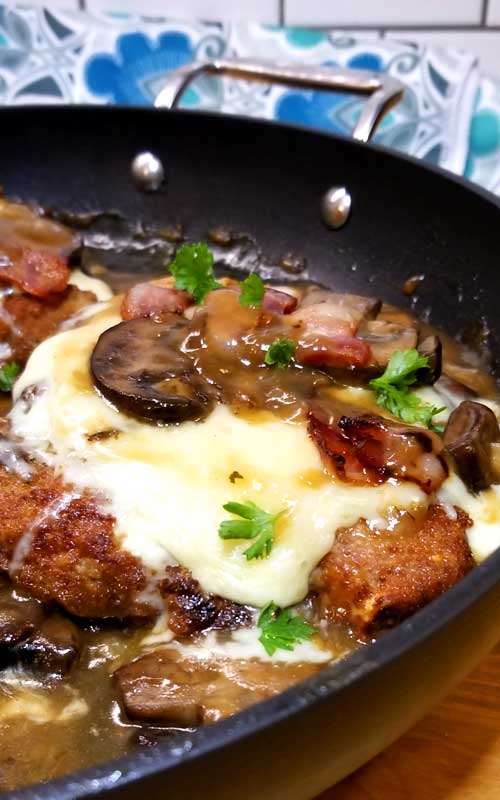 Close up view of a pork chop covered in Fontina cheese, mushroom gravy, mushrooms, and bacon. The pork chop is in a black skillet.