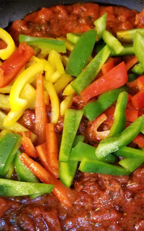 Uncooked red, yellow, and green peppers in a skillet, sitting on diced tomatoes.