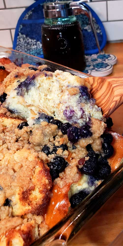 A prepared Blueberry French Toast Casserole with Streusel Topping being scooped from a glass baking dish. A  jar of maple syrup is in the background.