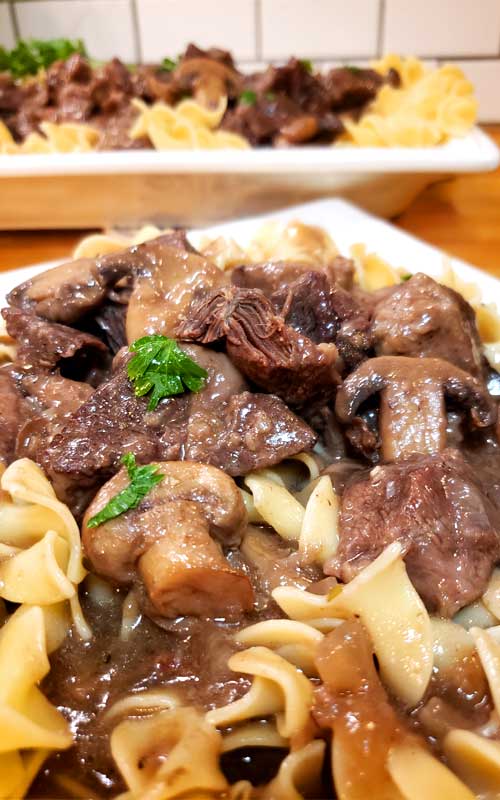 Close up view of Garlic Butter Beef Tips and Mushroom Gravy on a white plate, with a platter in the background holding more.