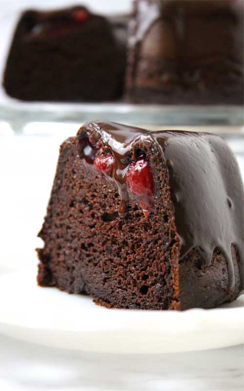 A slice of Chocolate Covered Cherry Bundt Cake on a white plate. The interior texture and cherry filling is visible. The cake is also covered in chocolate ganache. The rest of the cake can be seen in the background.