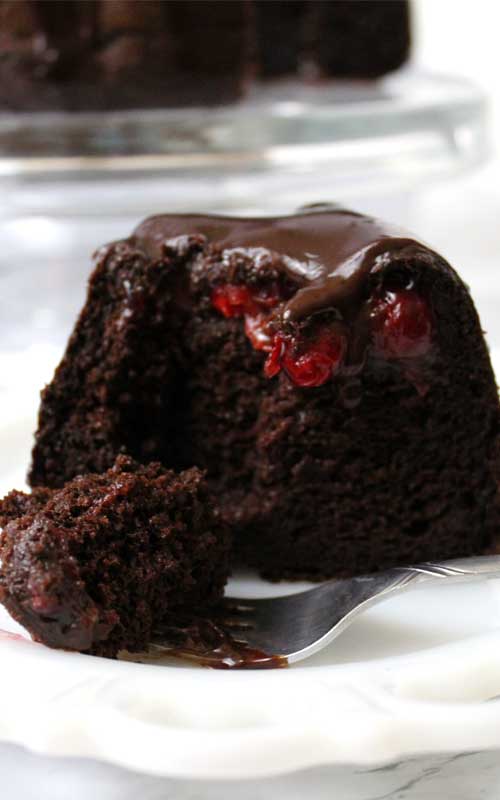 A slice of Chocolate Covered Cherry Bundt Cake on a white plate. The interior texture and cherry filling is visible. A part of the piece is on a silver fork in the foreground.
