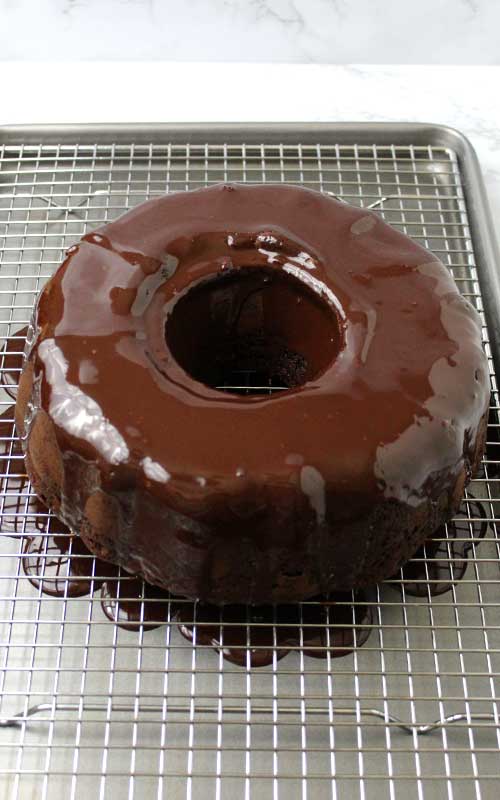 A whole Chocolate Covered Cherry Bundt Cake with chocolate ganache just poured over it. The cake is resting on a wire rack, with the excess ganache pooling up underneath.