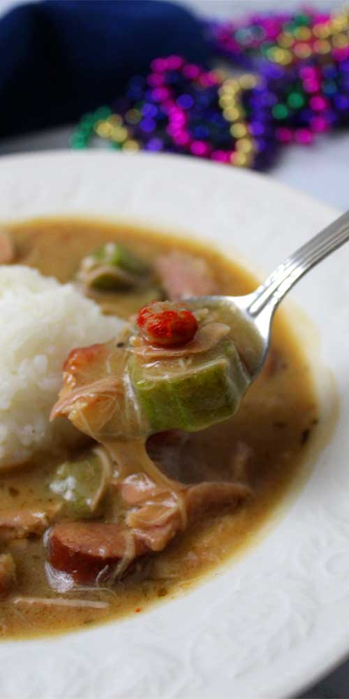 A white bowl filled with Authentic New Orleans Cajun Gumbo and white rice. A spoon has scooped out some okra, crawfish, and chicken.