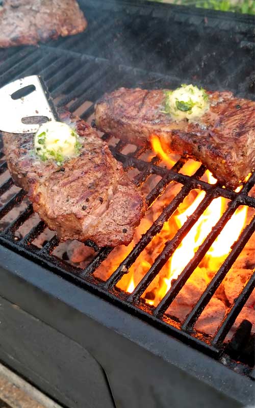Two Grilled Rib-Eye Steaks with Roasted Garlic Herb Butter being grilled on cast iron grates over charcoal that has flared up.