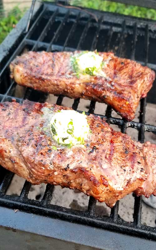Two Grilled Rib-Eye Steaks with Roasted Garlic Herb Butter resting on cast iron grates that are on a charcoal grill.