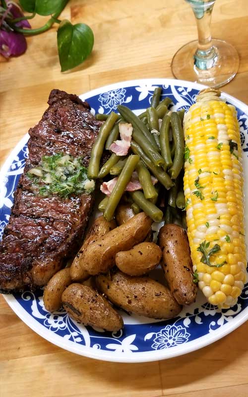 A Grilled Rib-Eye Steak with Roasted Garlic Herb Butter with green beans, an ear of corn, and fingerling potatoes on a blue and white plate.