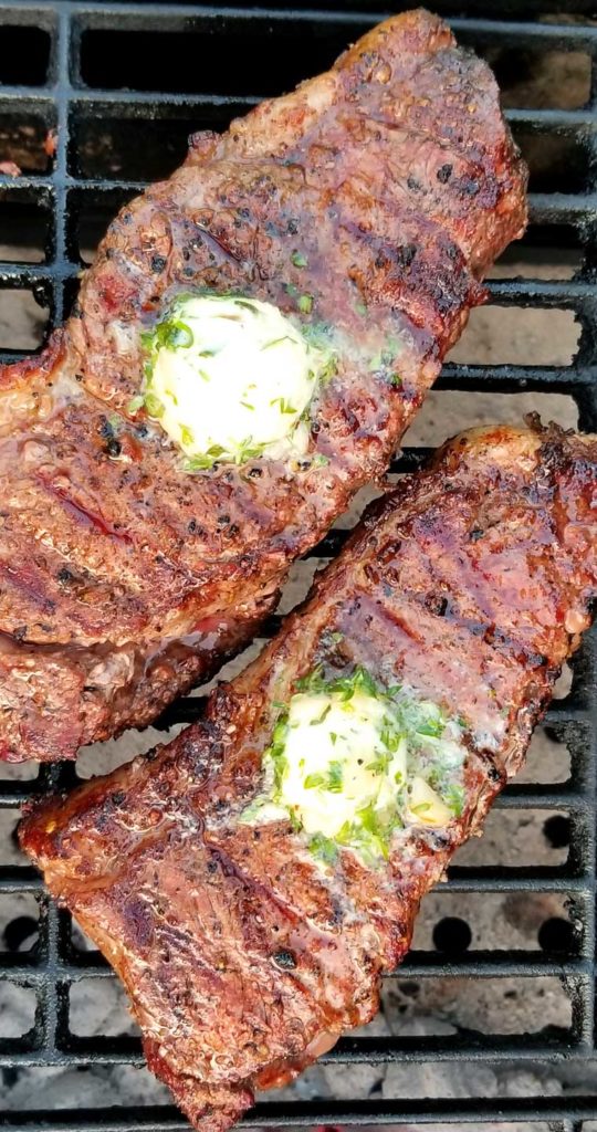 Two Grilled Rib-Eye Steaks with Roasted Garlic Herb Butter resting on a cast iron grate in a charcoal grill.