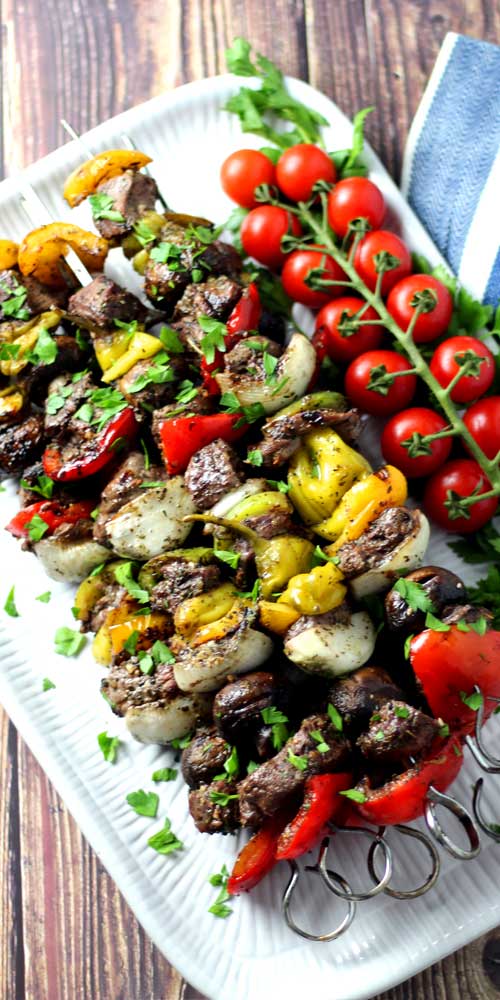 Five metal skewers going through pieces of steak, onion, pepperoncini, mushrooms, and red bell pepper. The food has been grilled, and is resting on a white platter. On the right side of the platter is a vine of red cherry tomatoes.