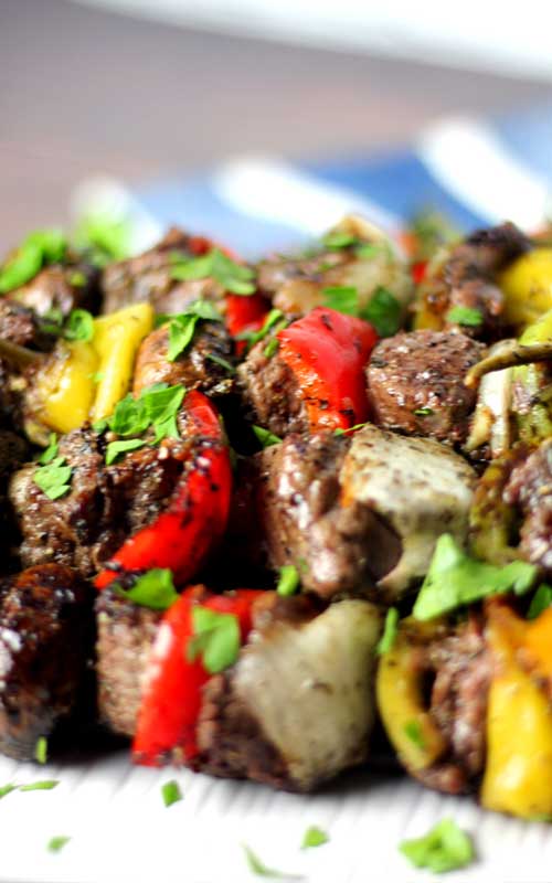 A close up view of prepared Italian Steak Kabobs. Steak, onion, and red bell pepper are clearly visible. The food is garnished with small pieces of torn parsley.