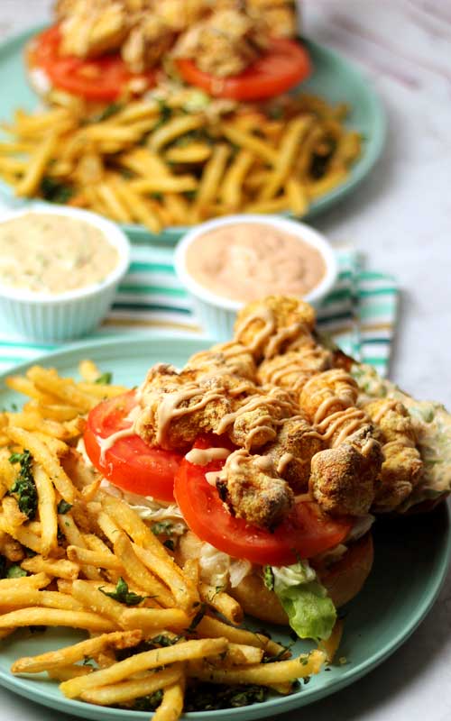 A Shrimp Po'Boy with Rémoulade Sauce, lettuce, and tomato on a blue plate. French fries are also on the plate. In the background is a similar plate, and two small cups of Rémoulade Sauce.