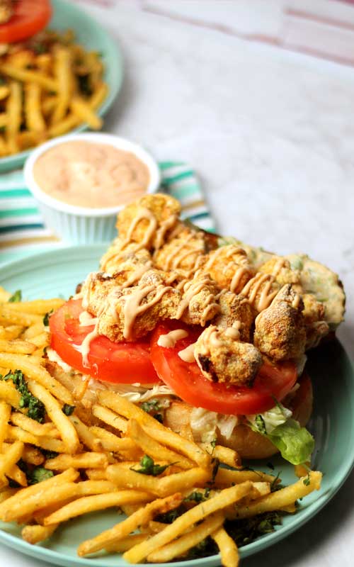 A Shrimp Po'Boy Rémoulade Sauce and French fries on a blue plate. A small cup of remoulade sauce and a similar plate can be seen in the background.