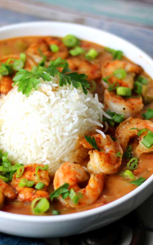 Shrimp in a tomato based gravy in a white bowl. In the center of the bowl is a mound of white rice. The dish is garnished with green onions and parsley.