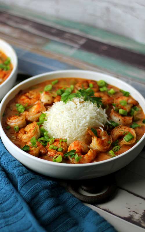 A white bowl filled with shrimp etouffee and white rice. The bowl is sitting on a multi-colored piece of wood, with a blue towel in the bottom-left corner.