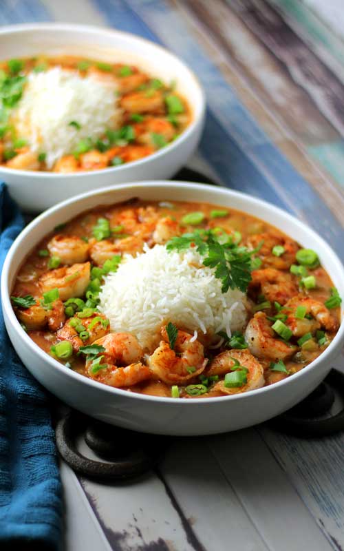 Two white bowl filled with large servings of shrimp etouffee. White rice, green onion, and parsley are also visible. The bowls are sitting on a multi-colored wood board.